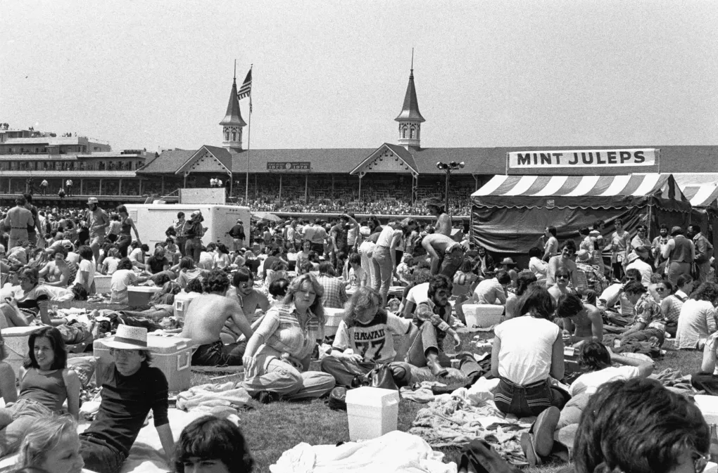A black and white photo of the infield crowd at the 1978 Kentucky Derby. The crowd is gathered around a tent selling mint juleps, with the historic Twin Spires behind it.