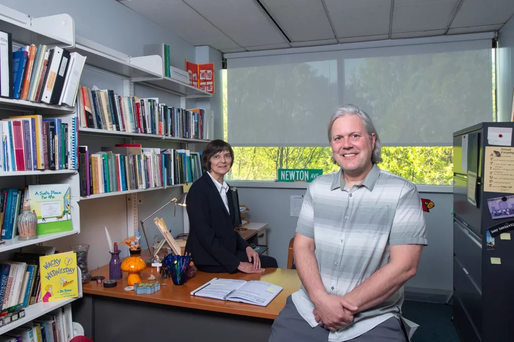 A woman and man pose on an office desk in front of a window.