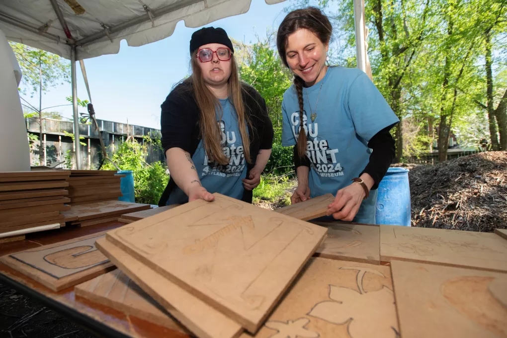 Two women work side by side together on detailed woodcuts at an outdoor art fair.