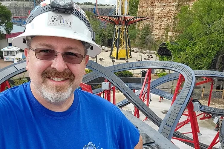 Alumnus Jeff Pike at an amusement park with a roller coaster behind him.