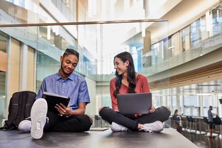 Two students are sitting on the floor while they study and do academic research on the second floor of the Belknap Academic Building.