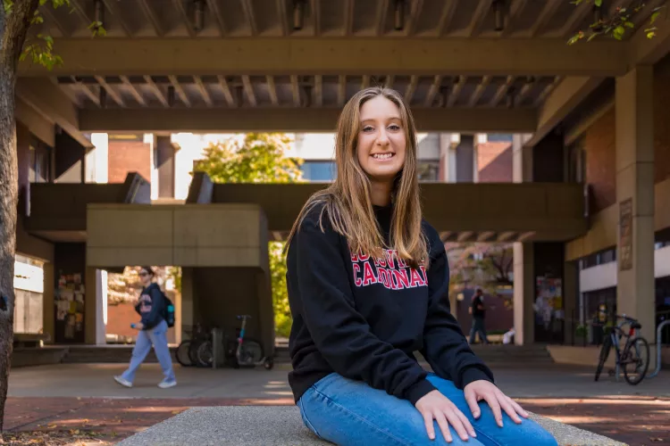 A young woman wearing a black sweatshirt with Louisville Cardinals in red lettering on the front poses for a picture on the UofL campus quad.