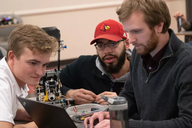 Three electrical and computer engineering students looking at a laptop alongside the small robot they made to compete in their class final.