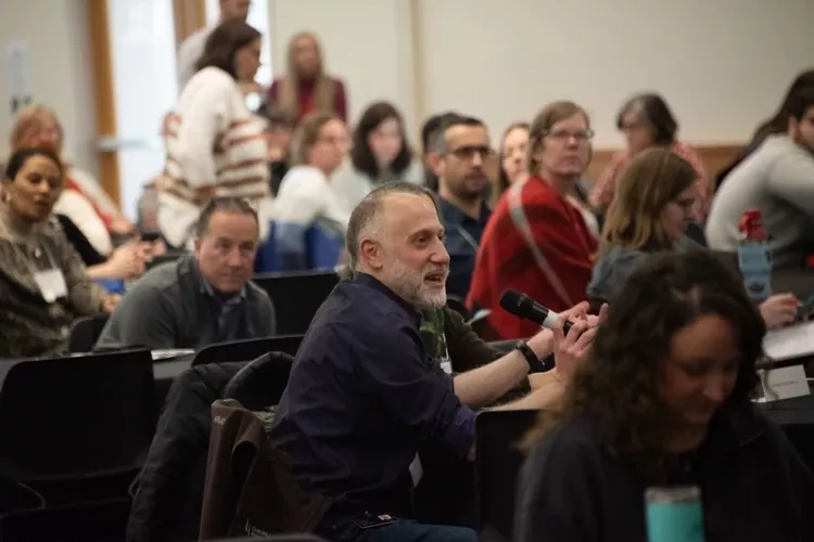 Participant seated at crowded table, speaking into microphone.