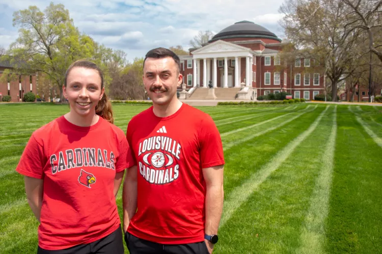 Two people wearing UofL t-shirts smile in a grassy lawn.