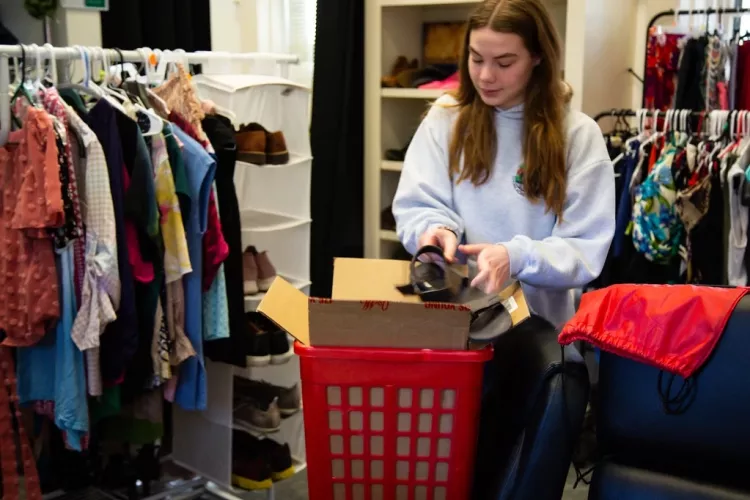 A person puts a shoe in a box, they are surrounded by clothing racks.