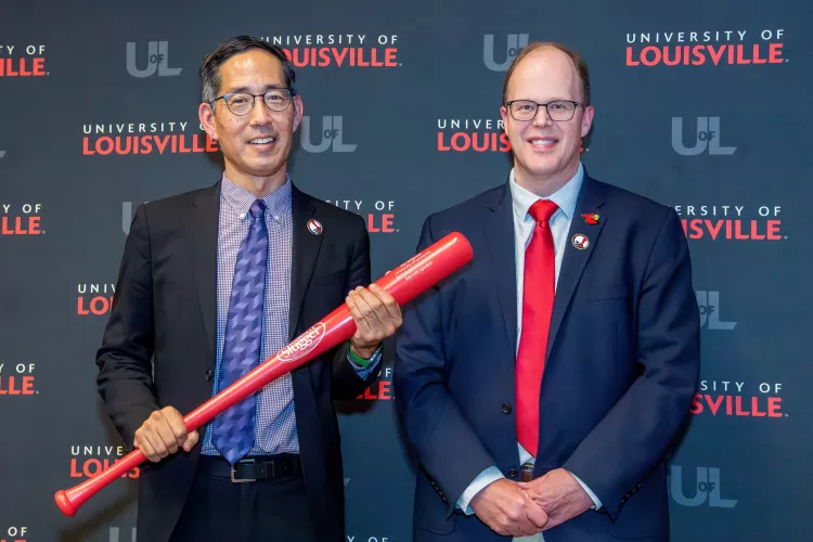 Two men in business suits, smiling. One holding a red baseball bat.