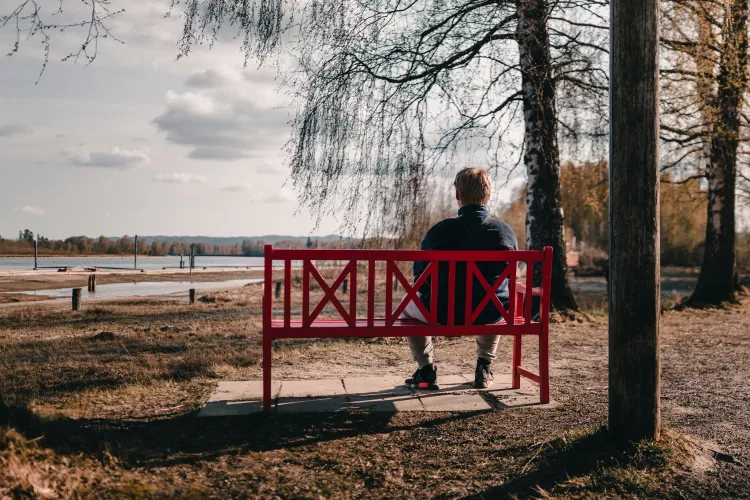 A man sits outside on a red bench, staring at a body of water.