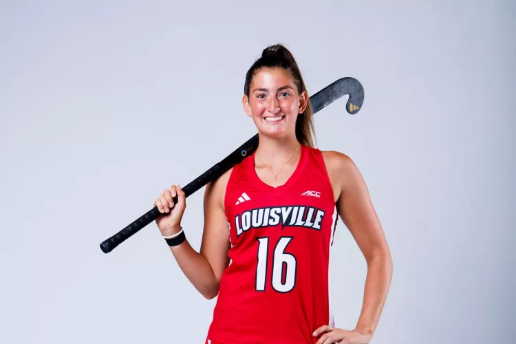 A portrait of UofL field hockey player Izzy Bianco in uniform holding a field hockey stick.