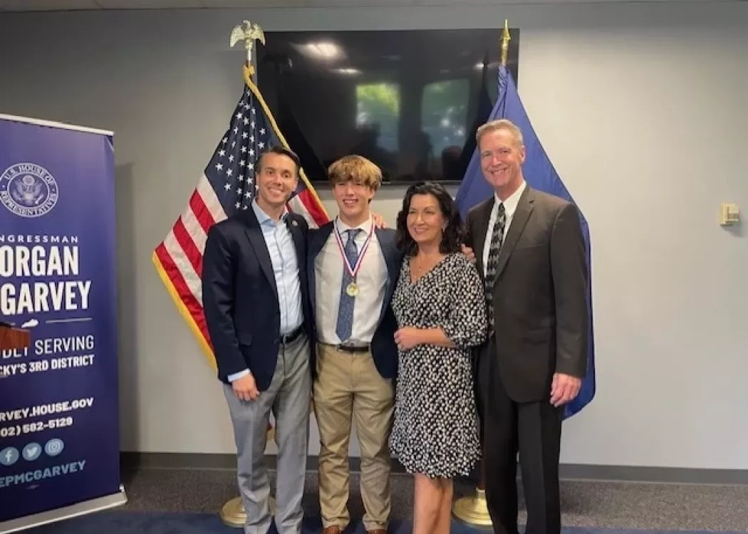 Congressman Morgan McGarvey, Jackson Robbins and Robbins' parents smile for a picture beside a United States flag and a Kentucky state flag.