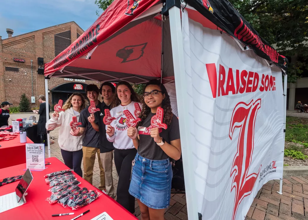 University of Louisville students holding foam hands in the shape of U L in front of a banner that reads "Raise Some L."