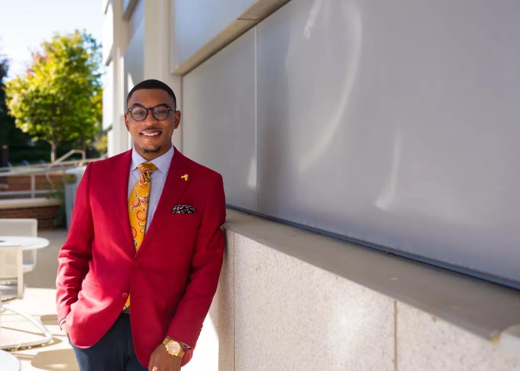 A young male sharply dressed in a red blazer and yellow tie poses for a photograph on UofL's campus.