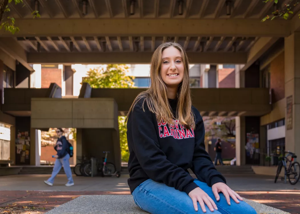 A young woman wearing a black sweatshirt with Louisville Cardinals in red lettering on the front poses for a picture on the UofL campus quad.