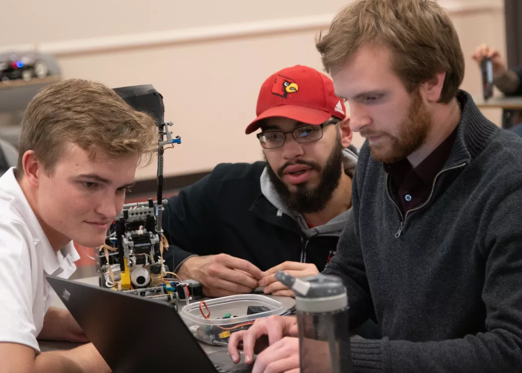 Three electrical and computer engineering students looking at a laptop alongside the small robot they made to compete in their class final.