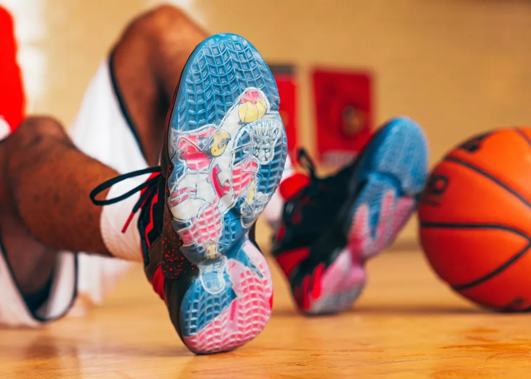 UofL Athletics Louie shoes close up with an image of Louie dunking a basketball. Basketball player wearing the shoes is sitting in the background with a basketball propped against their feet.