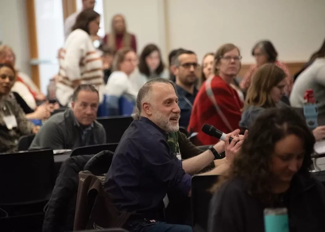 Participant seated at crowded table, speaking into microphone.