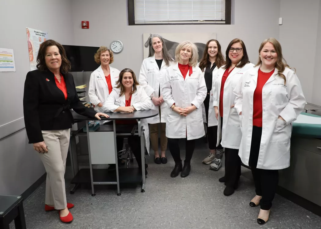 Kentucky Racing Health Services Center (KRHSC) Staff and University of Louisville School of Nursing faculty in an exam room located in the Center. A photo of a horse is behind the members. They are wearing their white lab coats.