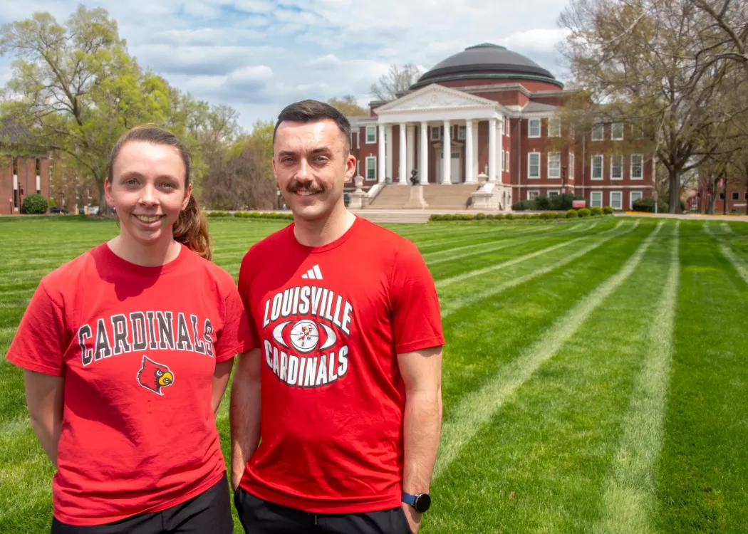 Two people wearing UofL t-shirts smile in a grassy lawn.