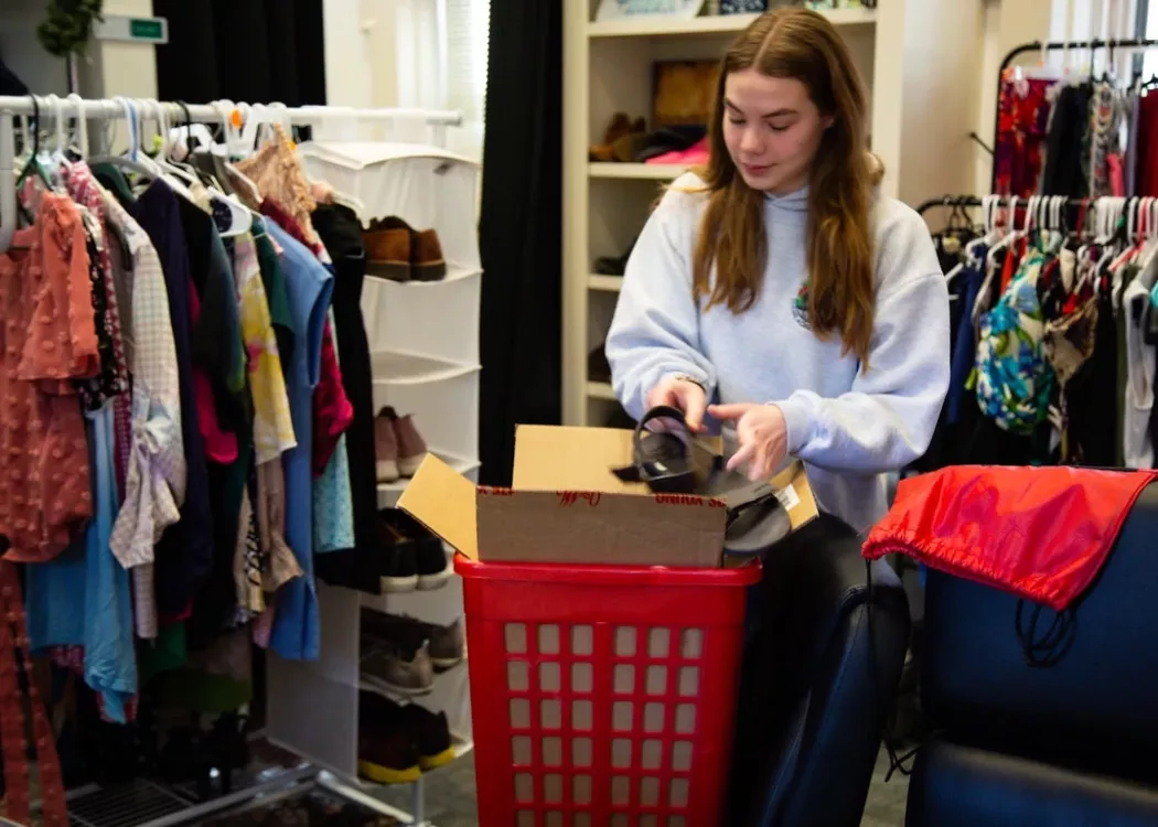 A person puts a shoe in a box, they are surrounded by clothing racks.