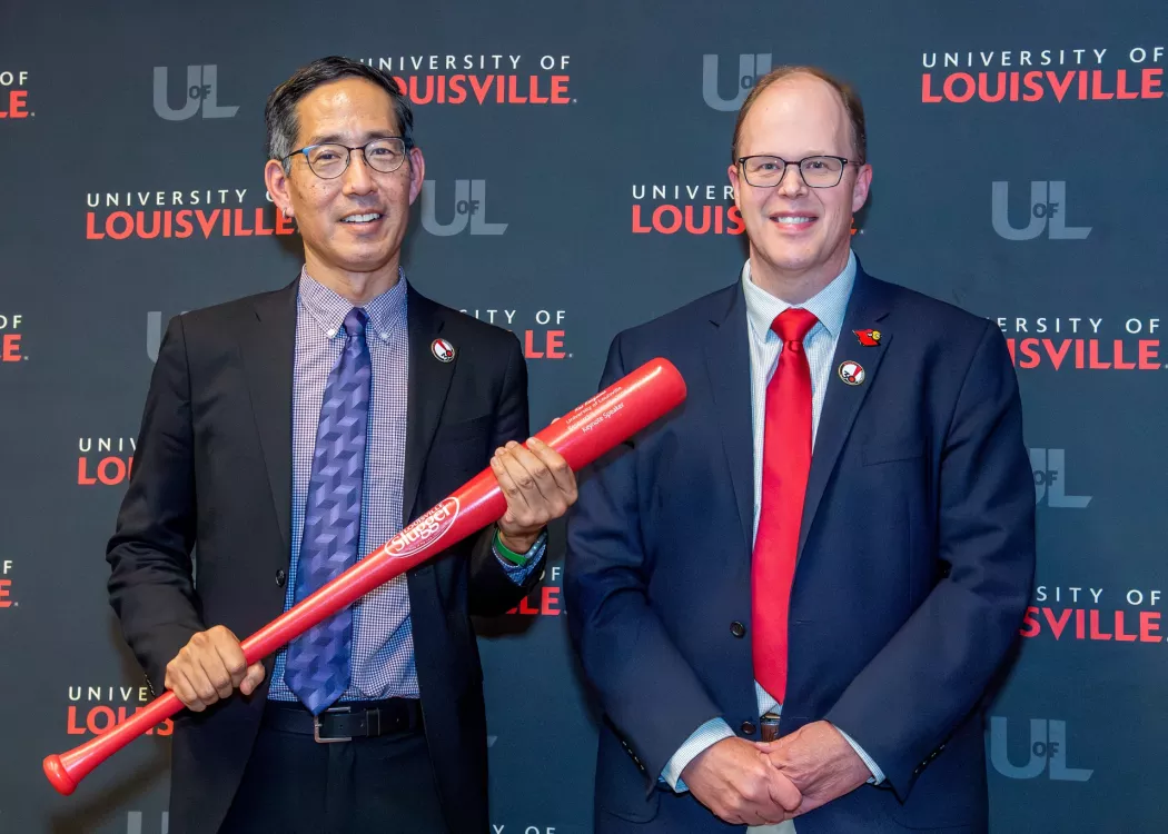 Two men in business suits, smiling. One holding a red baseball bat.