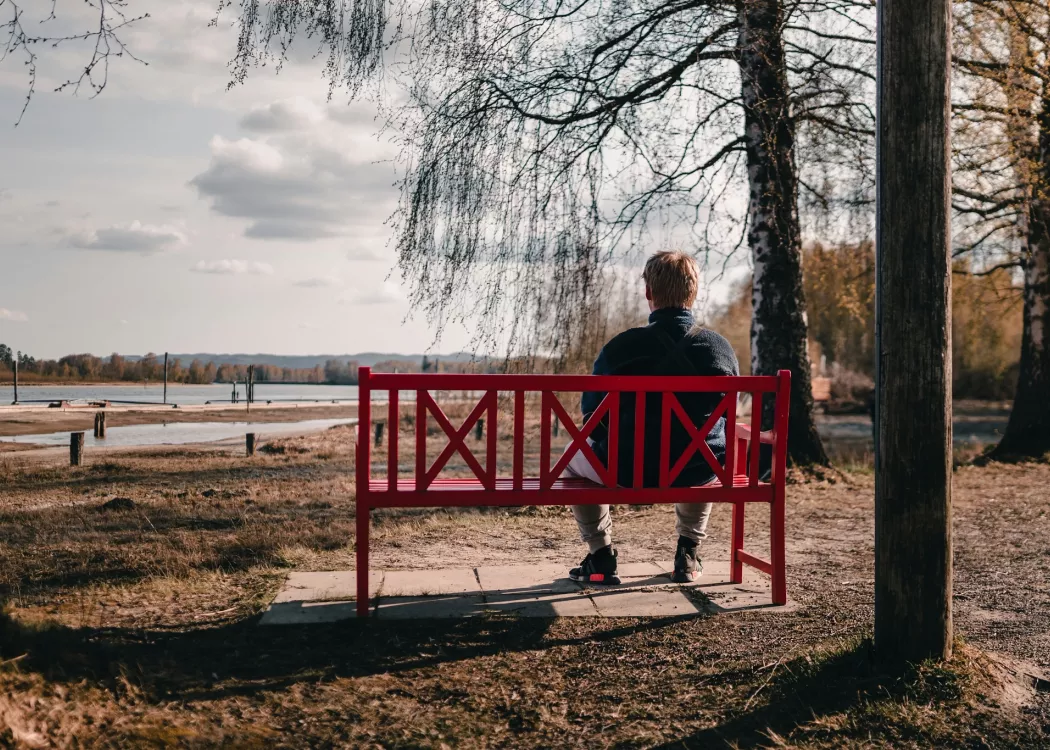 A man sits outside on a red bench, staring at a body of water.