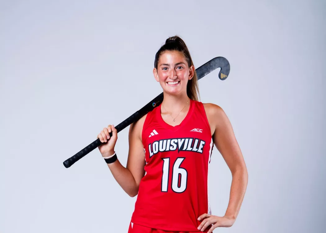 A portrait of UofL field hockey player Izzy Bianco in uniform holding a field hockey stick.