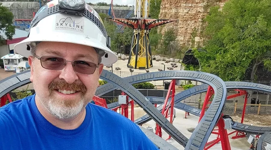 Alumnus Jeff Pike at an amusement park with a roller coaster behind him.