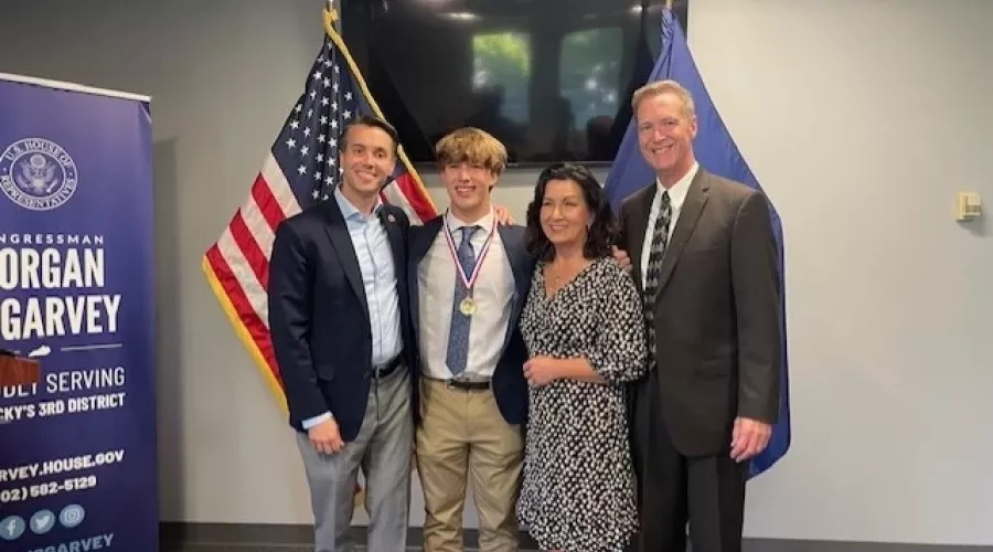 Congressman Morgan McGarvey, Jackson Robbins and Robbins' parents smile for a picture beside a United States flag and a Kentucky state flag.