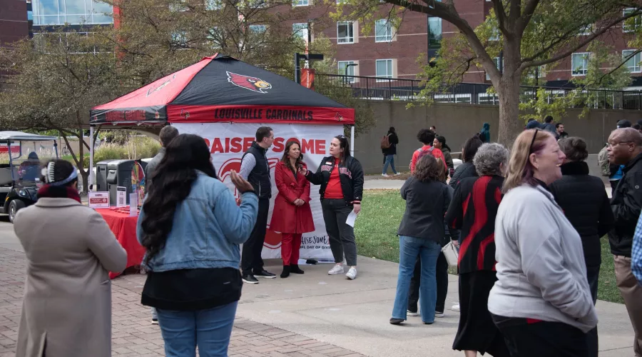 A crowd gathers around a woman in a University of Louisville Jacket interviewing two people.