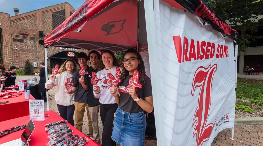 University of Louisville students holding foam hands in the shape of U L in front of a banner that reads "Raise Some L."