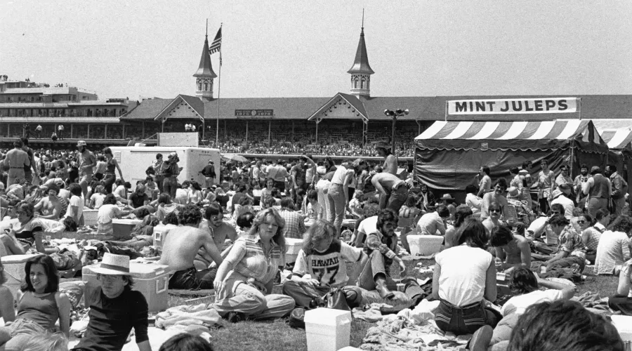 A black and white photo of the infield crowd at the 1978 Kentucky Derby. The crowd is gathered around a tent selling mint juleps, with the historic Twin Spires behind it.