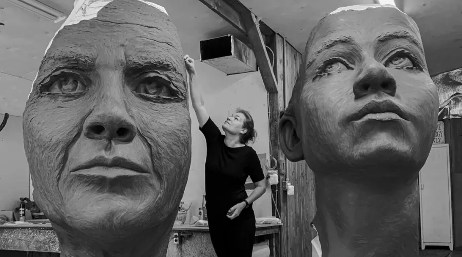 a woman is carving and surrounded by giant faces created out of clay in an artist studio.