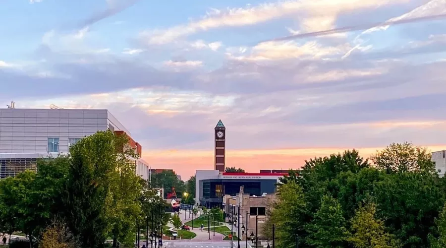 A sunrise over UofL creating a pink, yellow, and blue morning sky. UofL's clock tower and student union can be seen in the background amongst green trees.