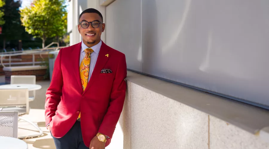 A young male sharply dressed in a red blazer and yellow tie poses for a photograph on UofL's campus.