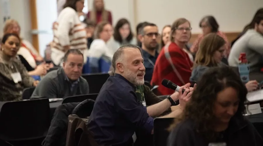 Participant seated at crowded table, speaking into microphone.
