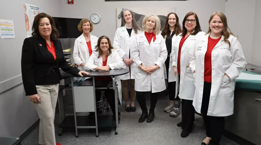 Kentucky Racing Health Services Center (KRHSC) Staff and University of Louisville School of Nursing faculty in an exam room located in the Center. A photo of a horse is behind the members. They are wearing their white lab coats.
