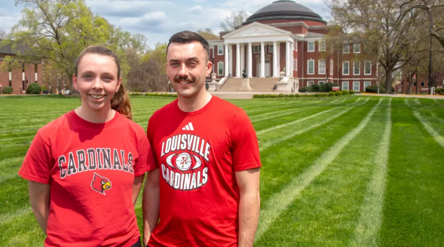Two people wearing UofL t-shirts smile in a grassy lawn.