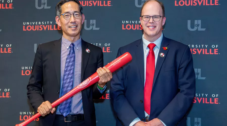 Two men in business suits, smiling. One holding a red baseball bat.