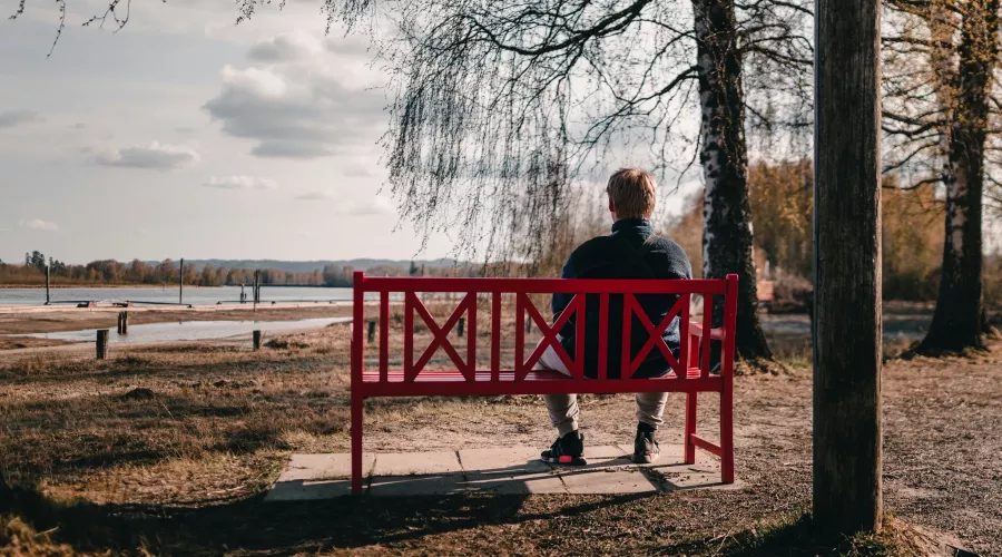 A man sits outside on a red bench, staring at a body of water.