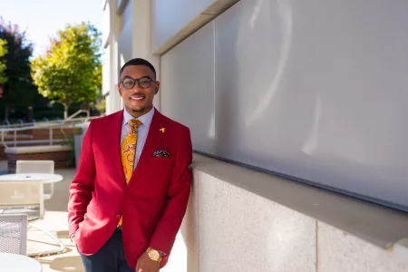 A young male sharply dressed in a red blazer and yellow tie poses for a photograph on UofL's campus.