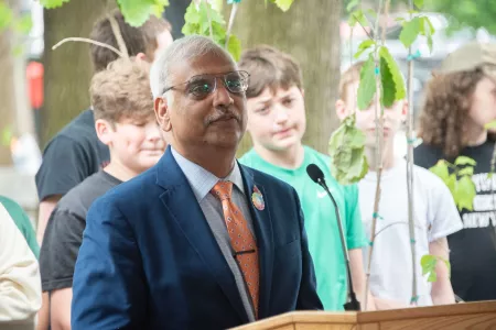 Man speaking at a podium with children and trees in background.