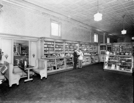 A black and white photograph of a pharmacy interior with a pharmacist talking to a customer in 1927.