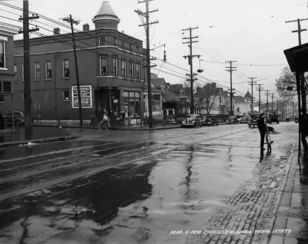 A black and white photograph of bicyclists and pedestrians meeting in the rain at a street corner in 1938.